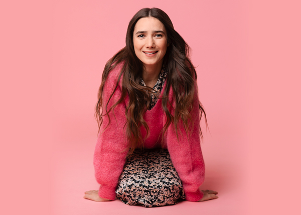 Linnea Leino on polvillaan maassa ja nojaa käsiinsä. Hänellä on päällä pörreä vaaleanpunainen neule ja kukallinen mekko.