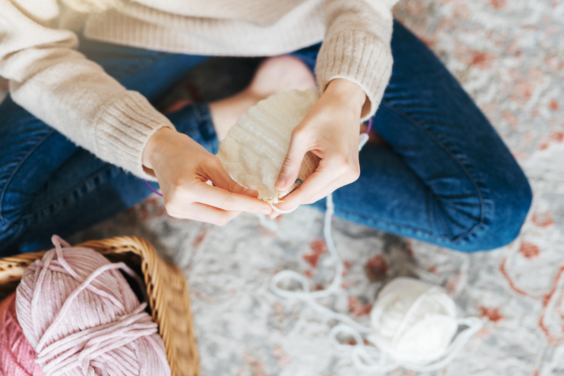 Nainen istuu jalat ristissä lattialla ja kutoo valkoisesta langasta kaulahuivia.