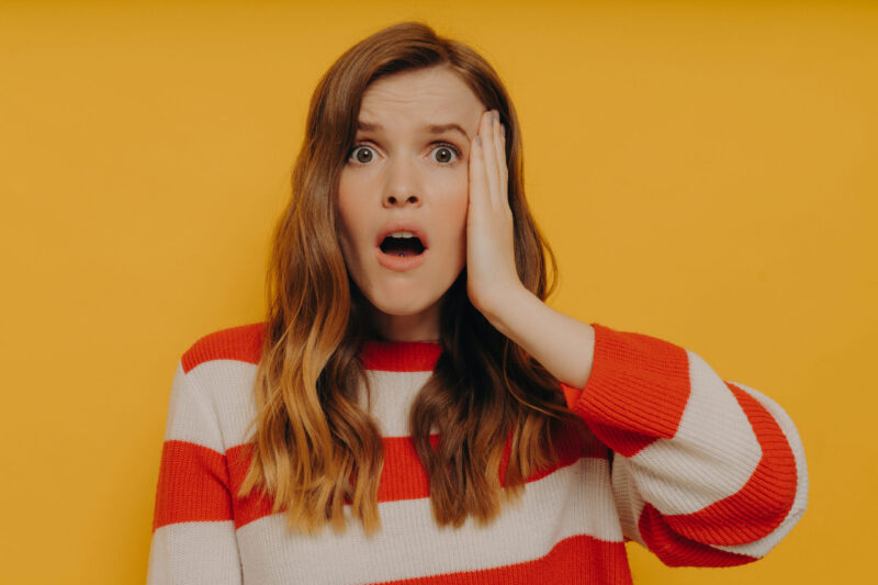 Nuori pitkähiuksinen kaunis nainen näyttää hämmentyneeltä. Hän pitelee toista kättä kasvoillaan suu auki.