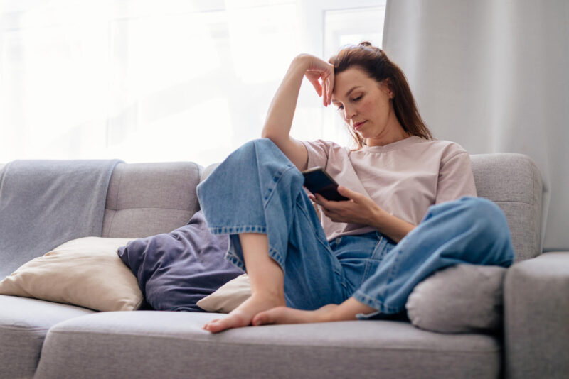 Nainen istuu sohvalla ja tuijottaa kännykkää.